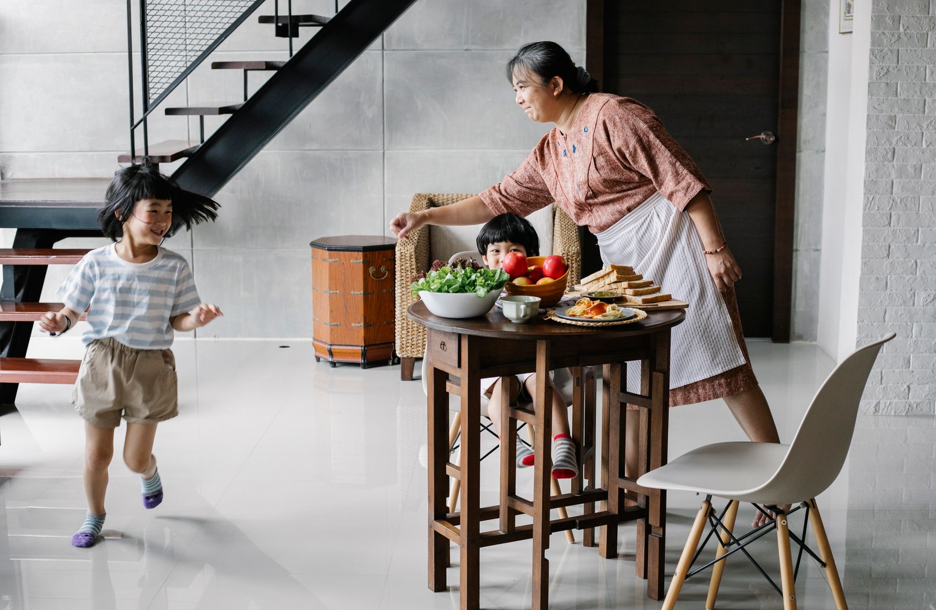 Full body of funny little Asian girl running around wooden table before having breakfast with brother and grandmother in stylish kitchen