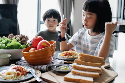 Serious little Asian girl eating delicious pasta with cute brother while sitting at table with healthy salad and apples on sunny day