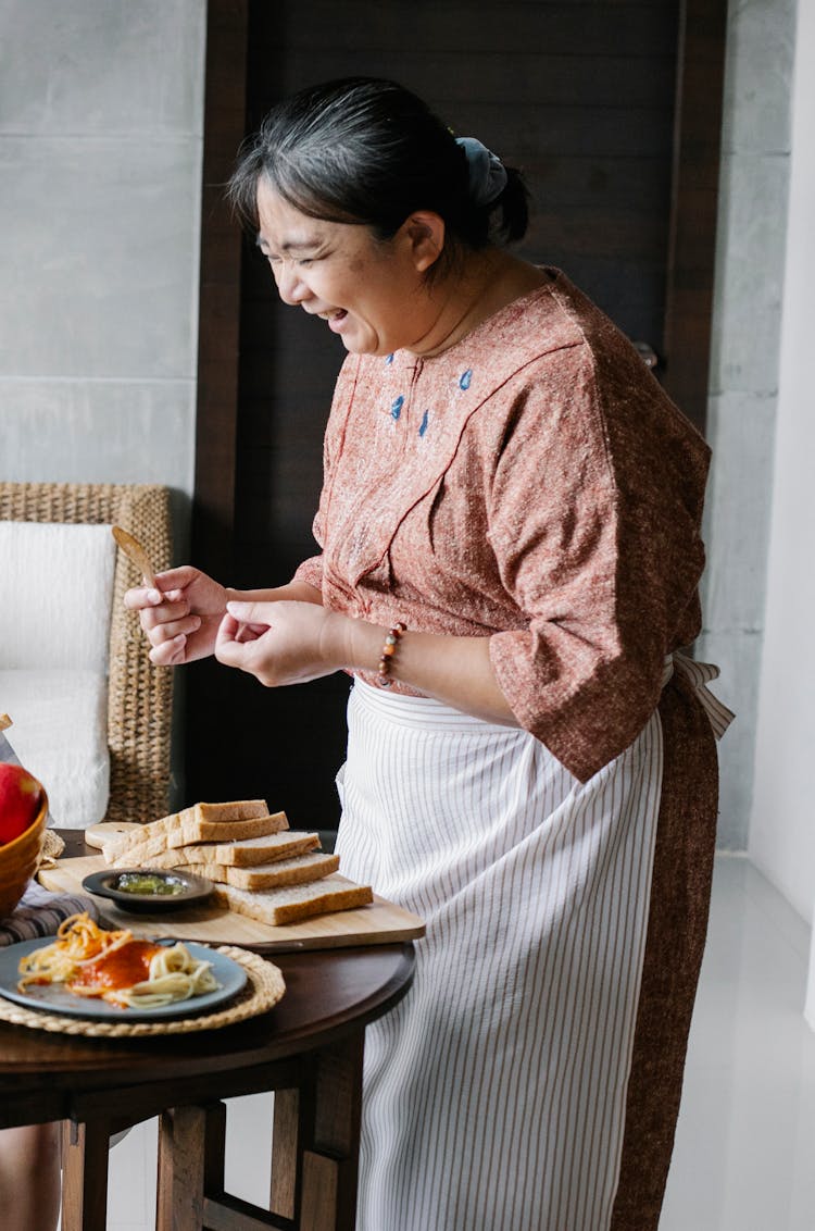 Cheerful Elderly Ethnic Female Laughing While Serving Lunch At Home