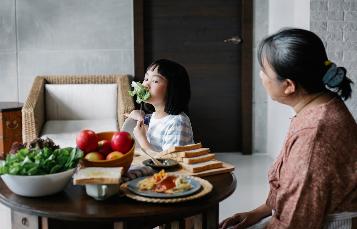 Little Asian girl eating healthy salad while sitting at table with grandmother