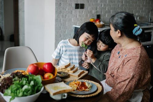 Cheerful little Asian siblings feeding each other while having lunch together with happy grandmother in modern kitchen