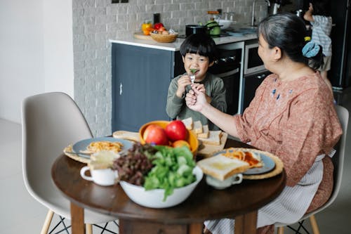 Cheerful little Asian boy having breakfast with kind grandmother at home