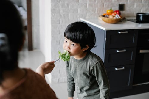Crop unrecognizable woman feeding cute little Asian son with healthy green lettuce leaves in modern kitchen