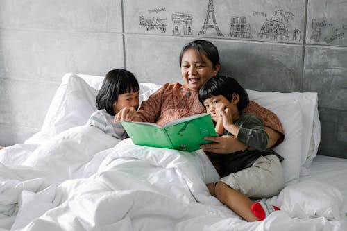 Free Adorable little Asian sibling lying on bed with smiling grandmother and reading book Stock Photo