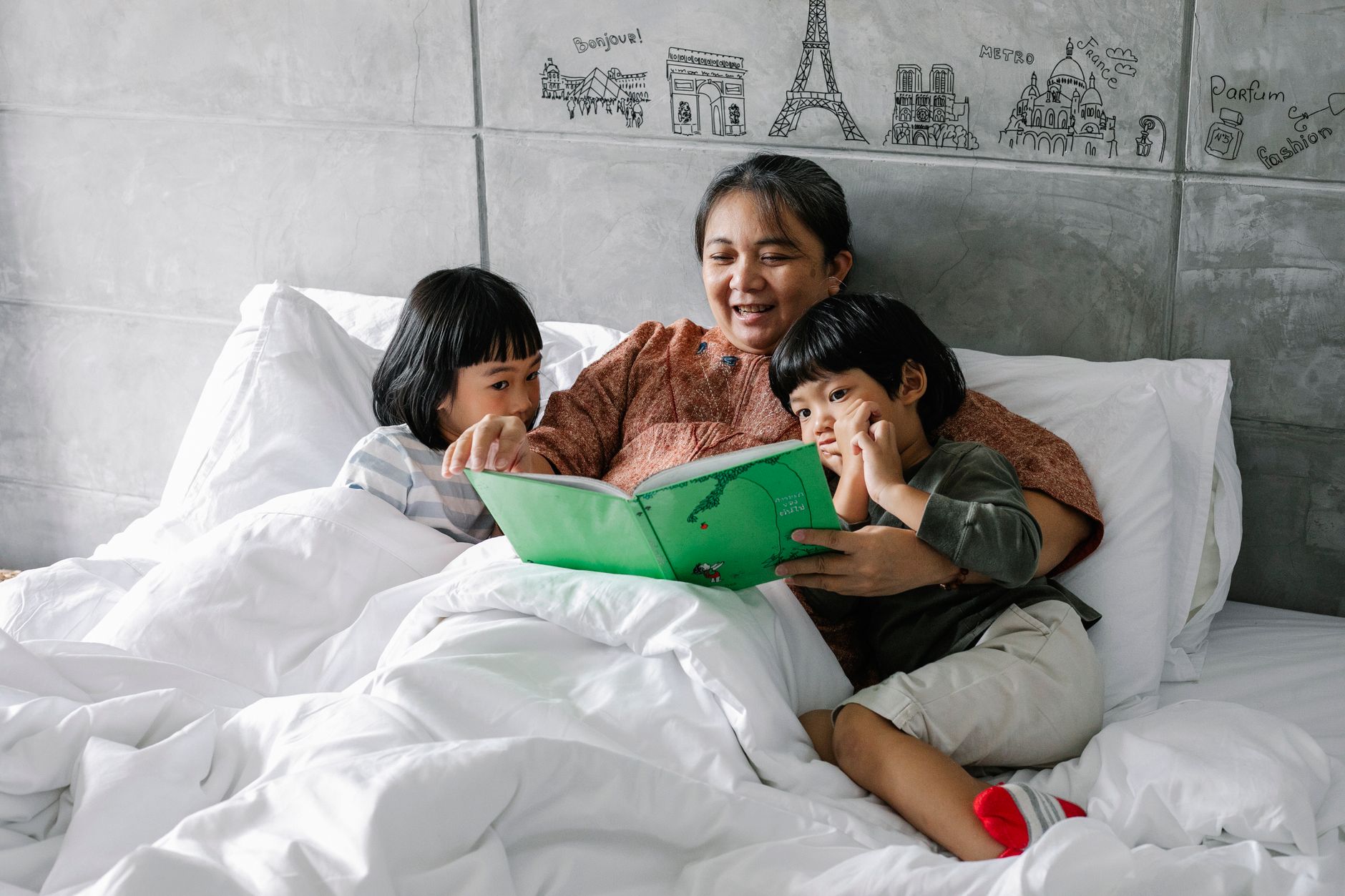 Positive aged ethnic grandmother smiling and reading book while lying on comfortable bed with cute attentive little grandchildren listening to story with interest