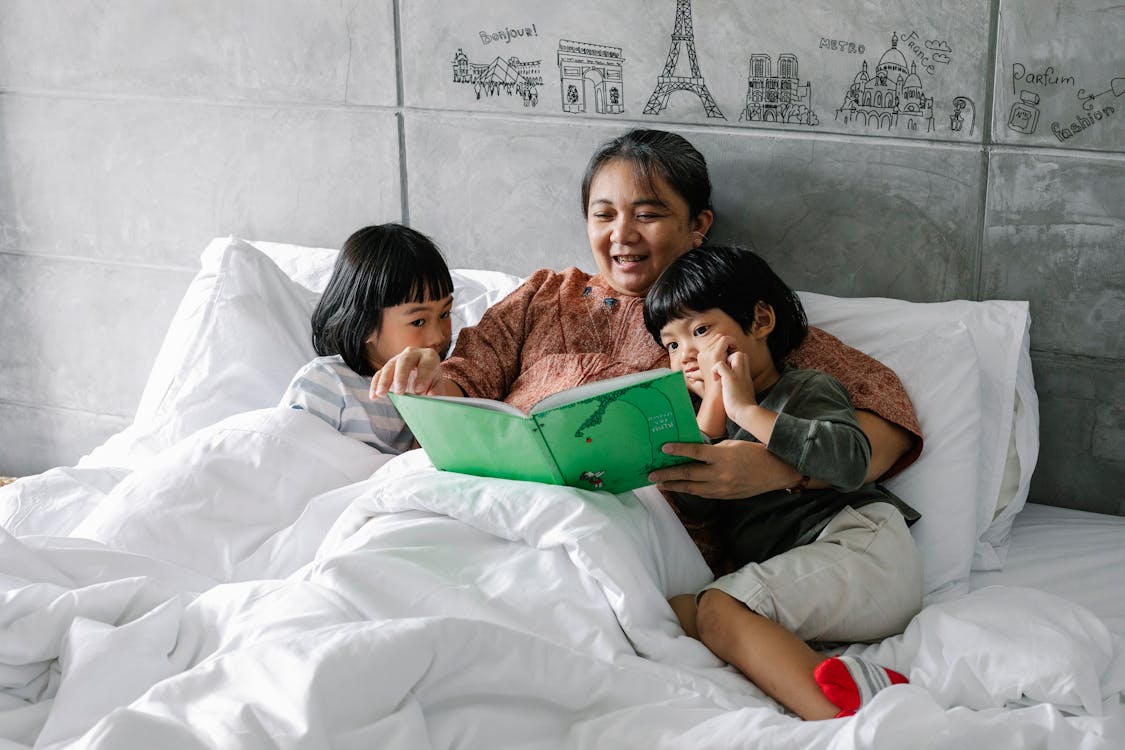 Adorable little Asian sibling lying on bed with smiling grandmother and reading book