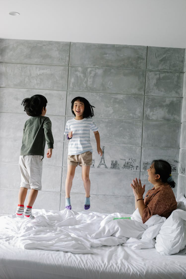 Excited Little Ethnic Children Jumping On Bed Near Clapping Grandmother