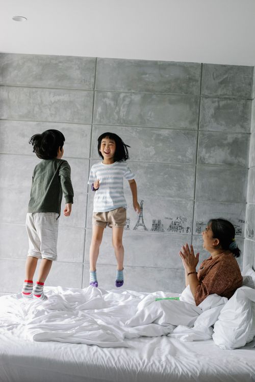 Free Adorable cheerful little Asian siblings jumping on bed and laughing while having fun together with happy grandmother at home Stock Photo