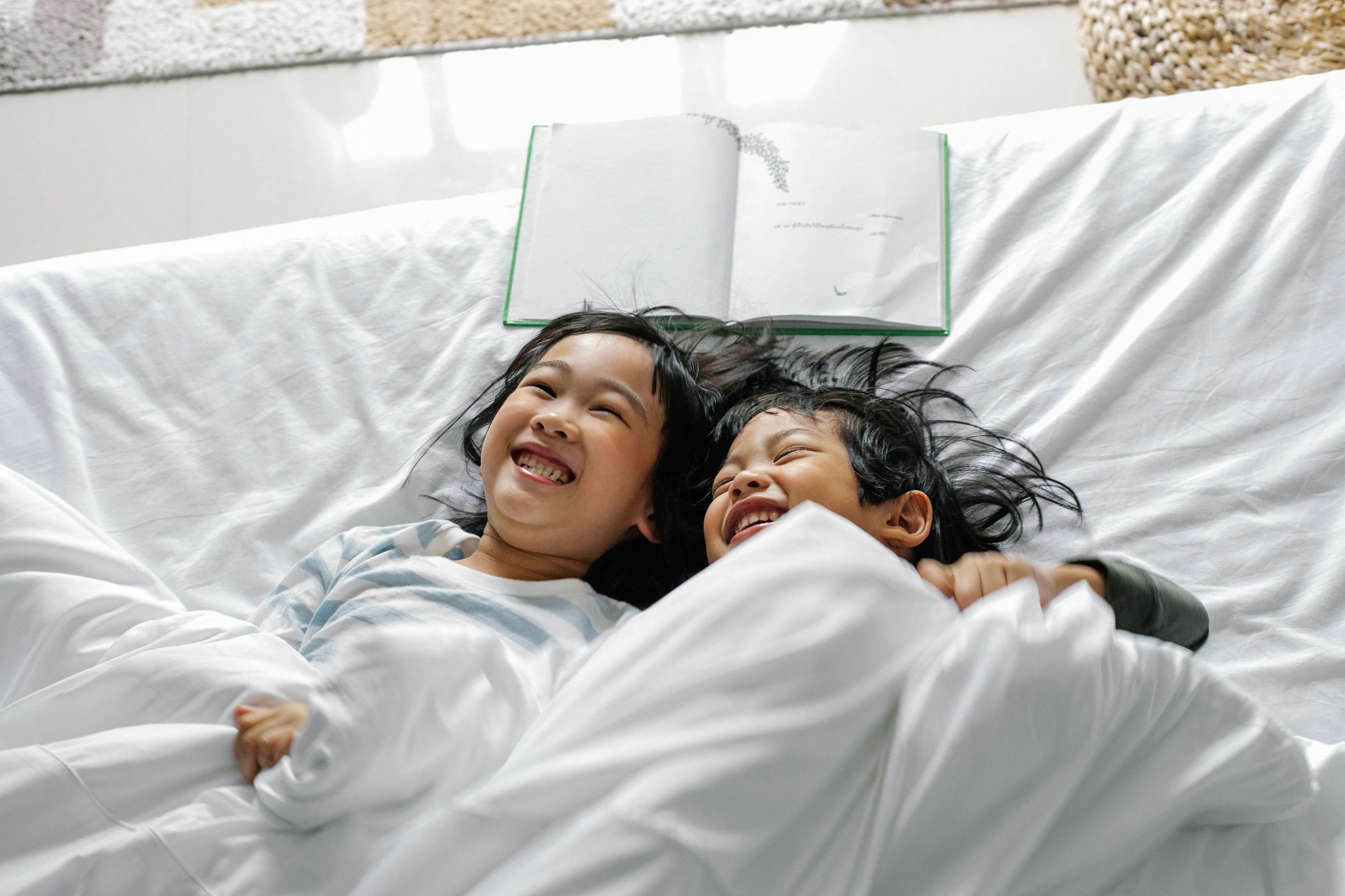 positive asian little siblings laughing while relaxing in bed