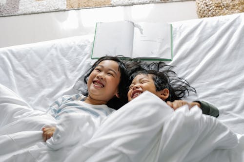 Positive Asian little siblings laughing while relaxing in bed