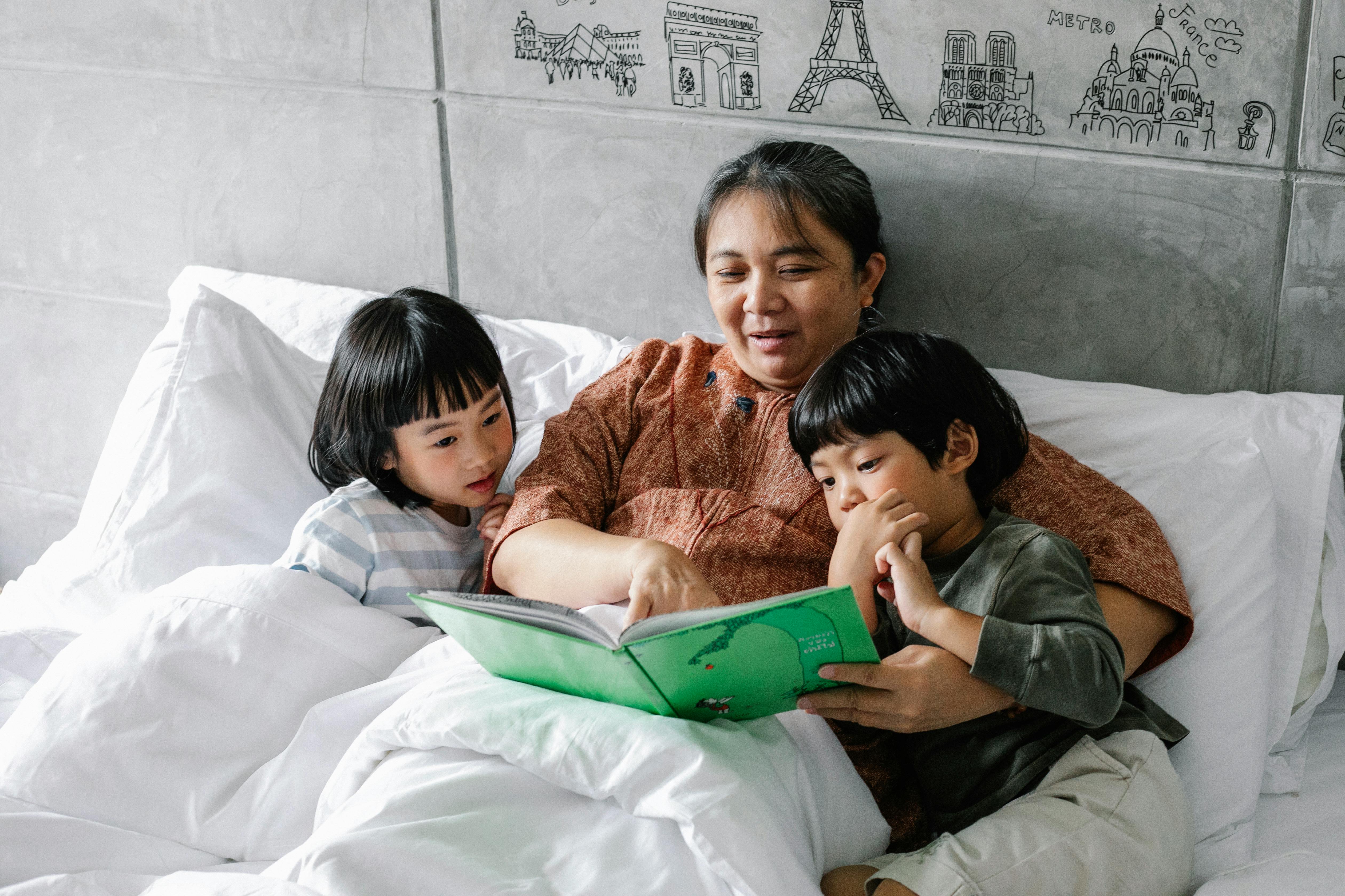 kind elderly ethnic woman lying in bed with curious grandchildren and reading book