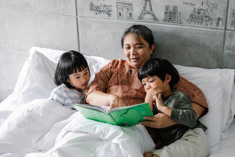 Kind Elderly Ethnic Woman Lying In Bed With Curious Grandchildren And Reading Book