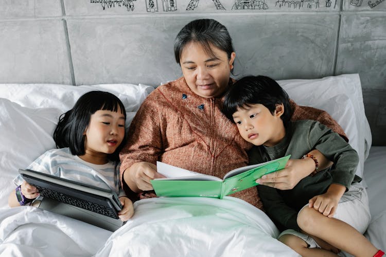 Ethnic Grandmother With Curious Children Lying On Bed With Interesting Book And Tablet