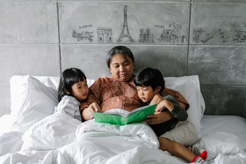 Free Serious Asian senior woman lying on bed and reading interesting story to attentive cute little siblings in stylish apartment Stock Photo