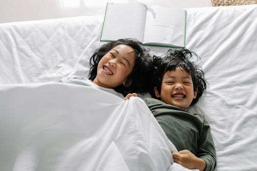 Free From above of Asian siblings laughing happily while lying in bed under soft blanket Stock Photo