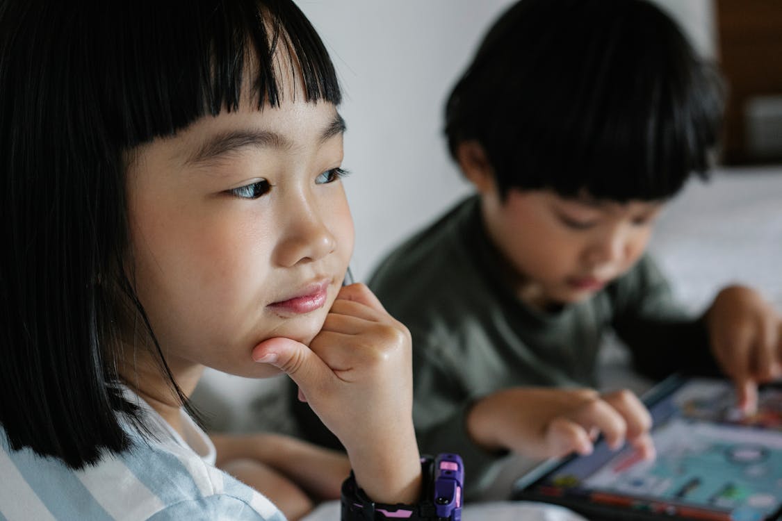 Free Crop concentrated ethnic girl touching chin while sitting near sibling playing game in modern device Stock Photo