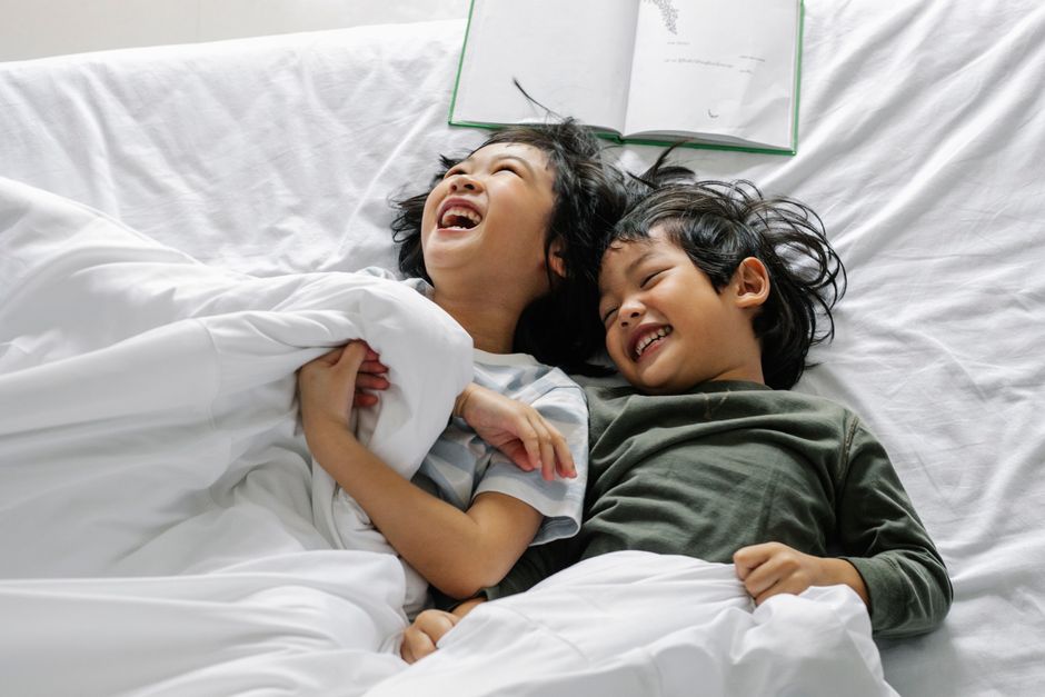 From above of happy Asian siblings having fun in cozy bed together during daytime sleep