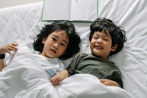 Free From above of happy Asian boy and girl lying on bed with book under blanket and looking at camera in bedroom Stock Photo