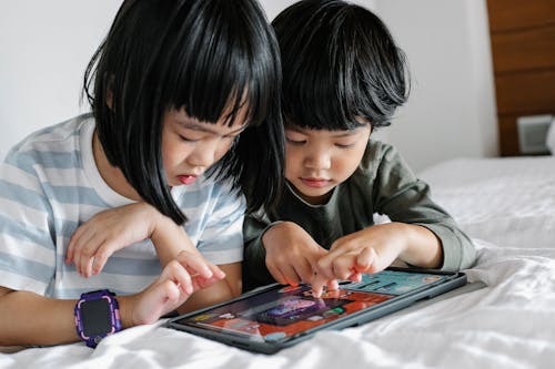 Attentive Asian boy and girl playing video game on modern tablet while lying on bed in bedroom in morning time