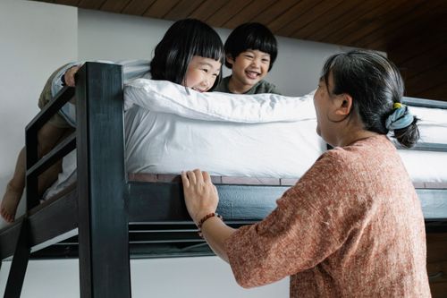 Free Side view of grandmother standing near bunk bed with cheerful Asian boy and girl in morning time in light bedroom Stock Photo