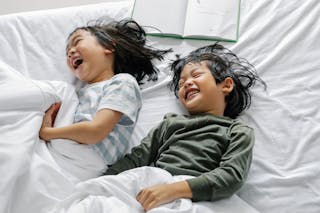 From above of Asian boy and girl laughing and smiling while lying in sleepwear on bed in morning n bedroom