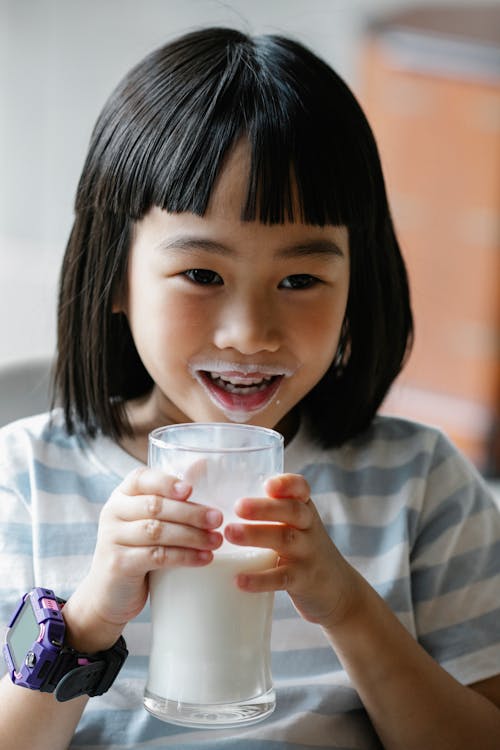 Positive Asian preteen with milk on lips and glass of healthy beverage in hands looking away while sitting in kitchen