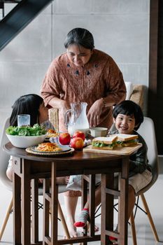 祖母坐在厨房里吃早餐，餐桌旁供应着三明治、牛奶和水果，她身旁站着亚洲男孩和女孩