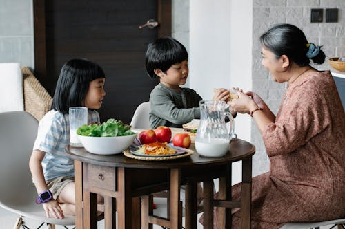 Asian boy and girl sitting at served table with fruits and milk with grandmother during breakfast time in morning on kitchen