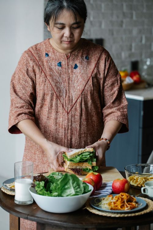 Free Serious Asian middle aged female in casual dress making sandwich with fresh lettuce leaves while standing near table in kitchen Stock Photo
