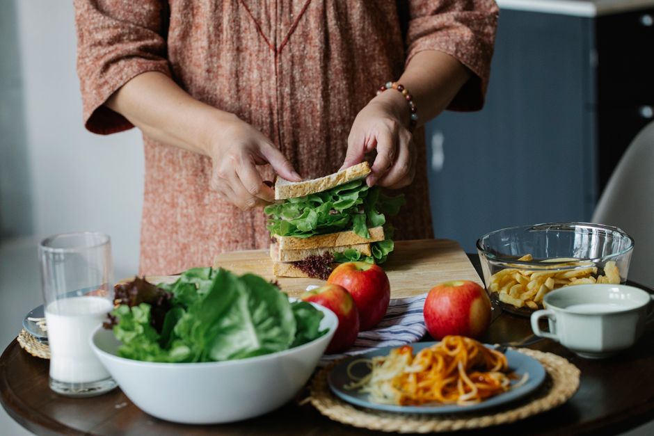 Unrecognizable female cook putting slice of bread on green lettuce while standing at table with assorted food and making breakfast