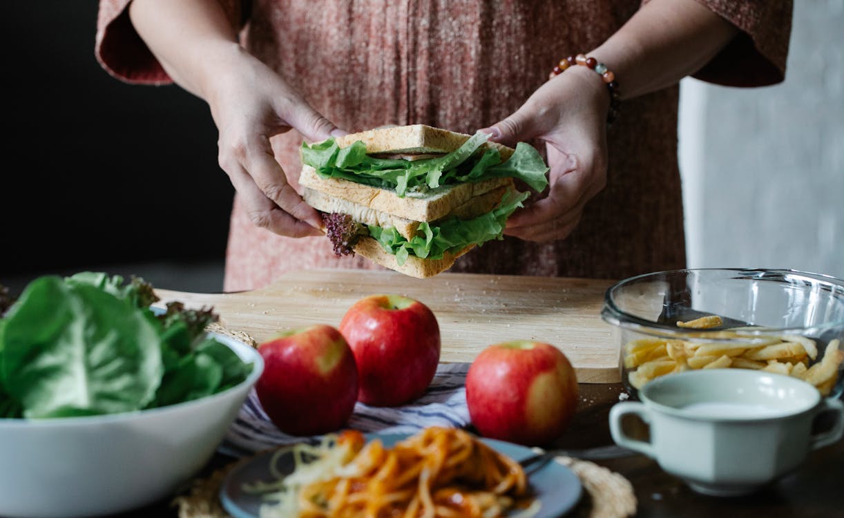 Free Unrecognizable female standing at table with apples and cutting board with sandwich with lettuce in hands in kitchen on breakfast time Stock Photo