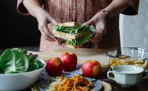 Unrecognizable female standing at table with apples and cutting board with sandwich with lettuce in hands in kitchen on breakfast time