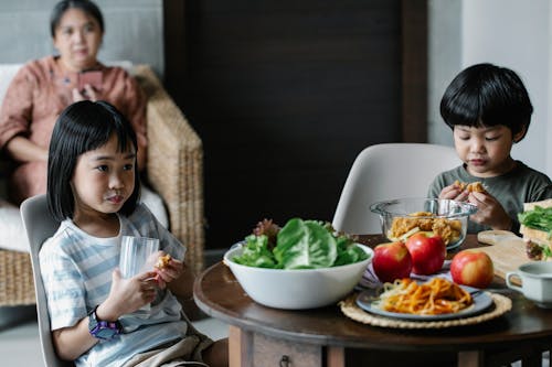 Calm Asian boy and girl sitting at served table while drinking milk during breakfast in kitchen with blurred grandmother on background