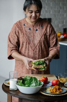 Content Asian mature female in casual cloth standing at table with sandwich with lettuce while making breakfast in kitchen in morning