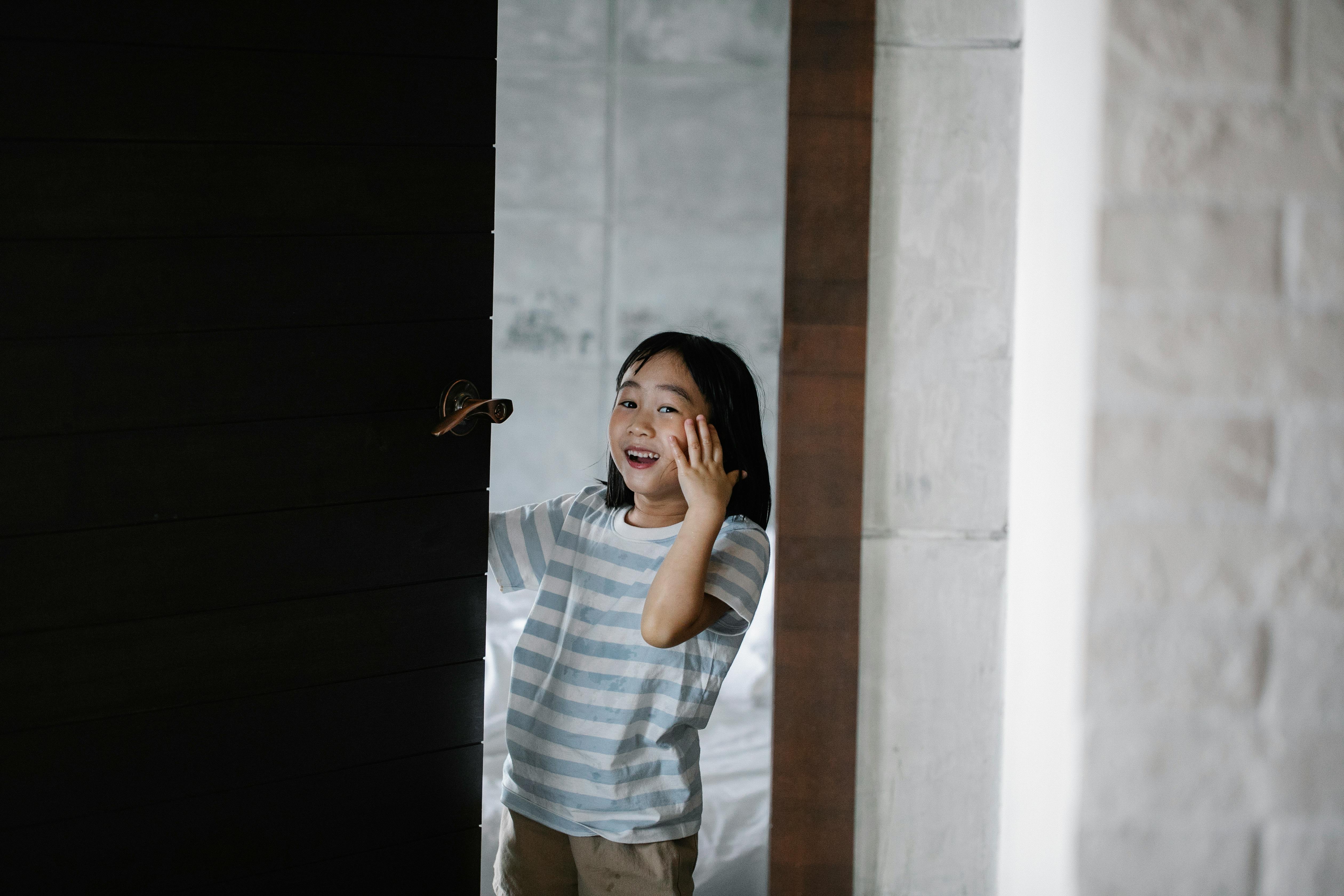 Cheerful Asian kid looking at camera while standing near wooden door and white brick wall with hand on cheek in flat
