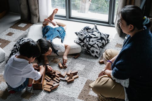 From above of Asian boy and girl playing with wooden blocks near window on floor in living room with grandmother