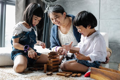 Free Happy grandmother sitting on floor with Asian boy and girl while playing with wooden blocks and building tower in living room near window Stock Photo