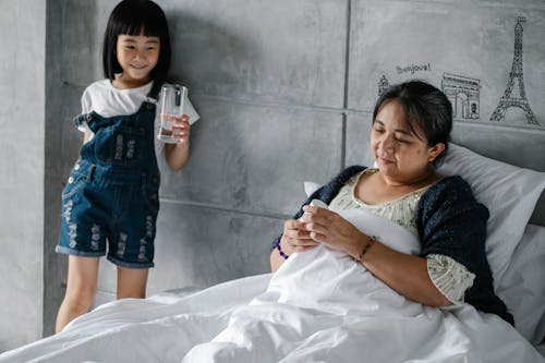 Adorable Asian little girl holding glass of water while standing near senior ill grandmother lying in bed in morning