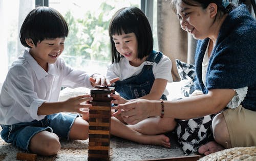 Full body of happy ethnic little children with elderly grandmother sitting on floor and constructing wooden tower while playing board game at home