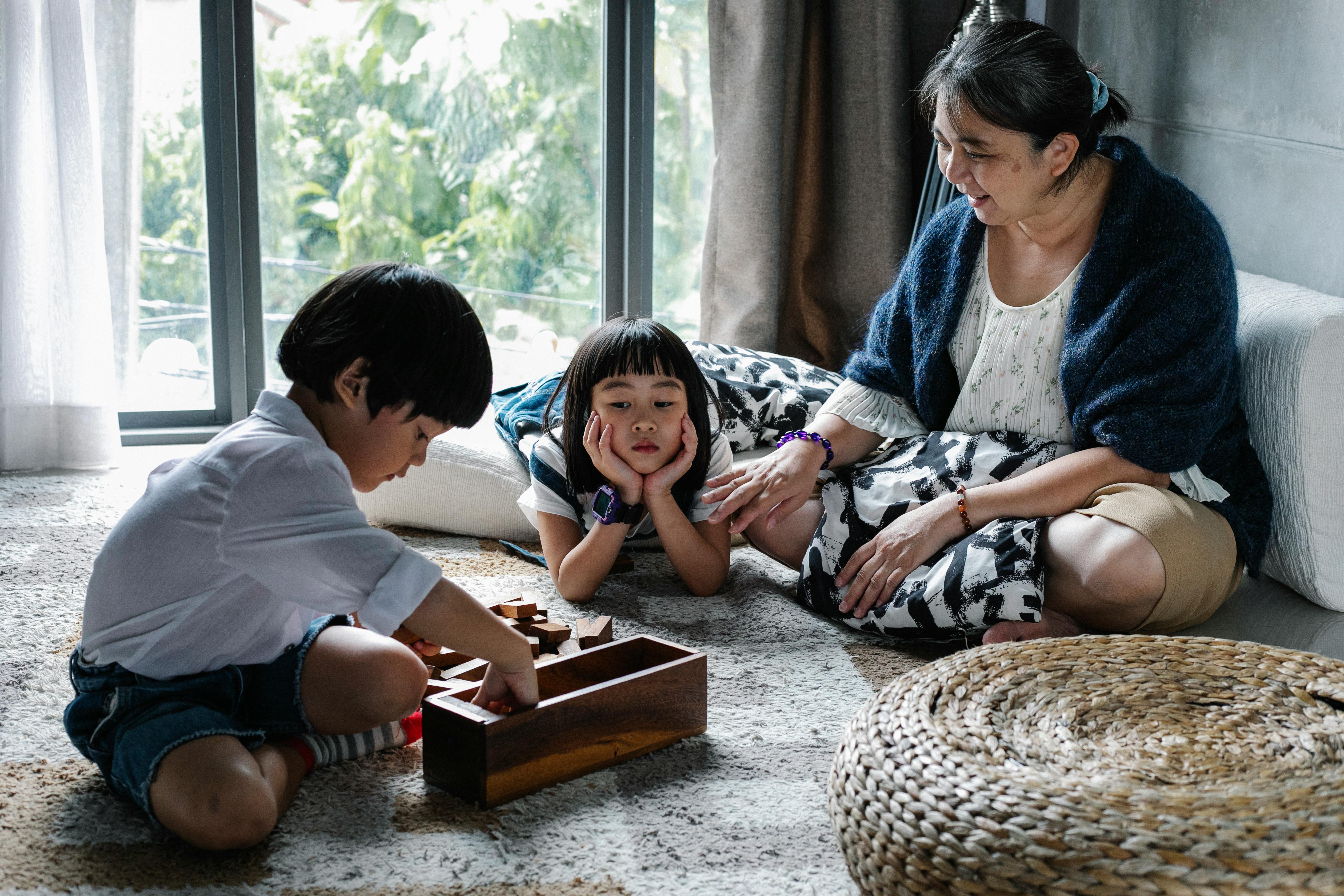 upset ethnic kid looking at brother picking up blocks after playing game with grandmother