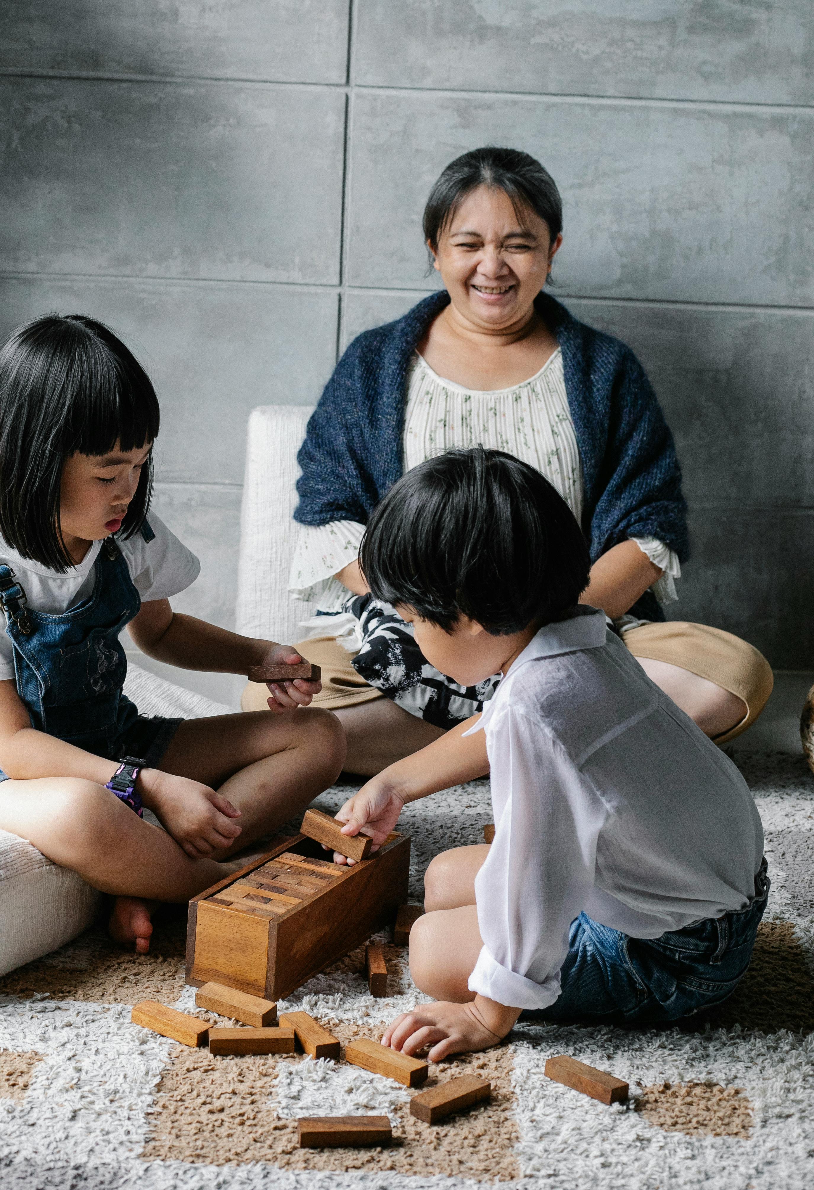 concentrated asian children playing tower game sitting on floor near cheerful grandmother