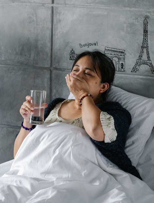Elderly sick Asian female lying in comfortable bed and drinking pill with glass of water