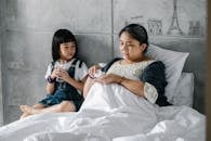 Focused little Asian girl sitting on bed near sick grandmother drinking pills