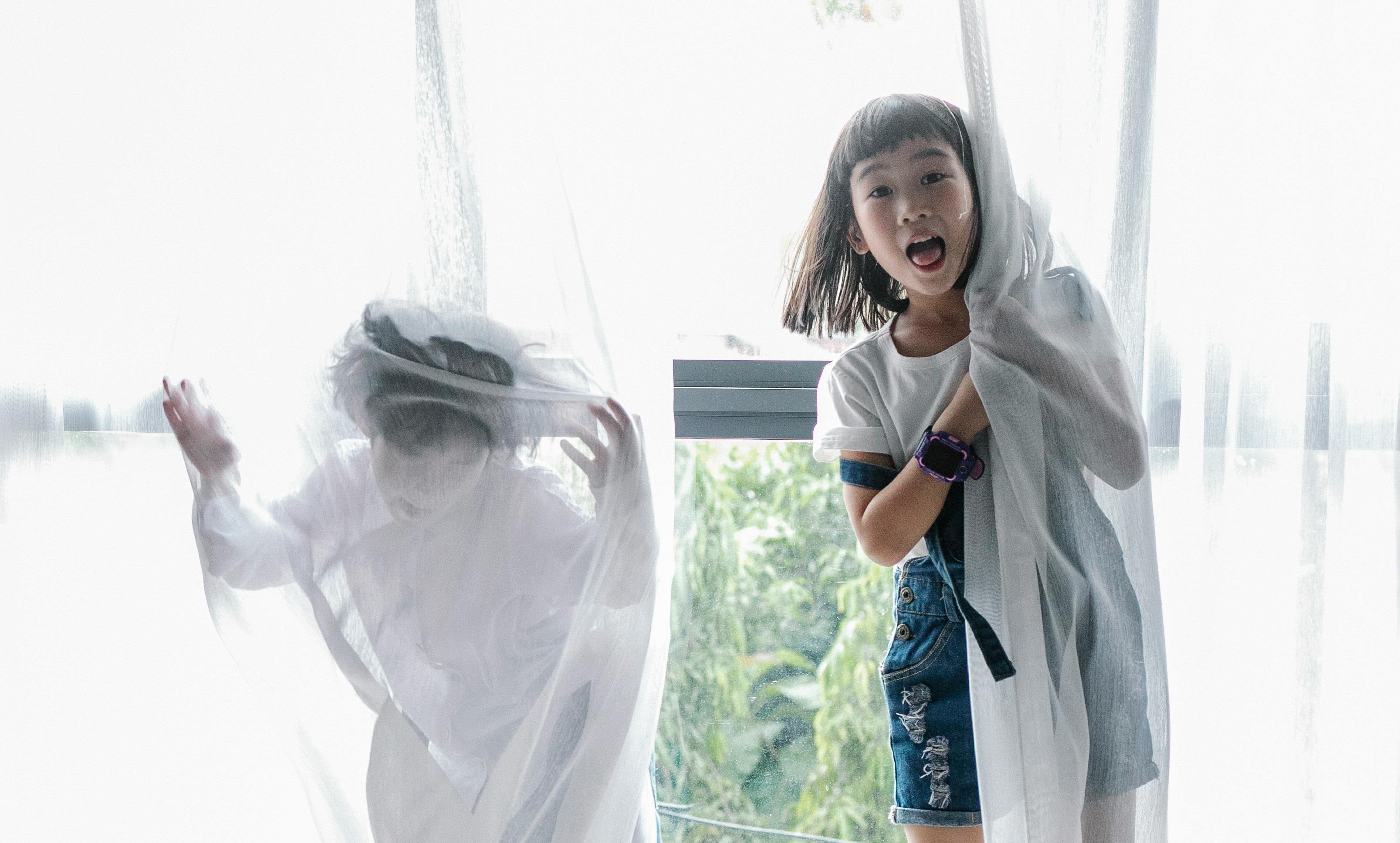 funny ethnic children hiding behind curtain while playing together at home