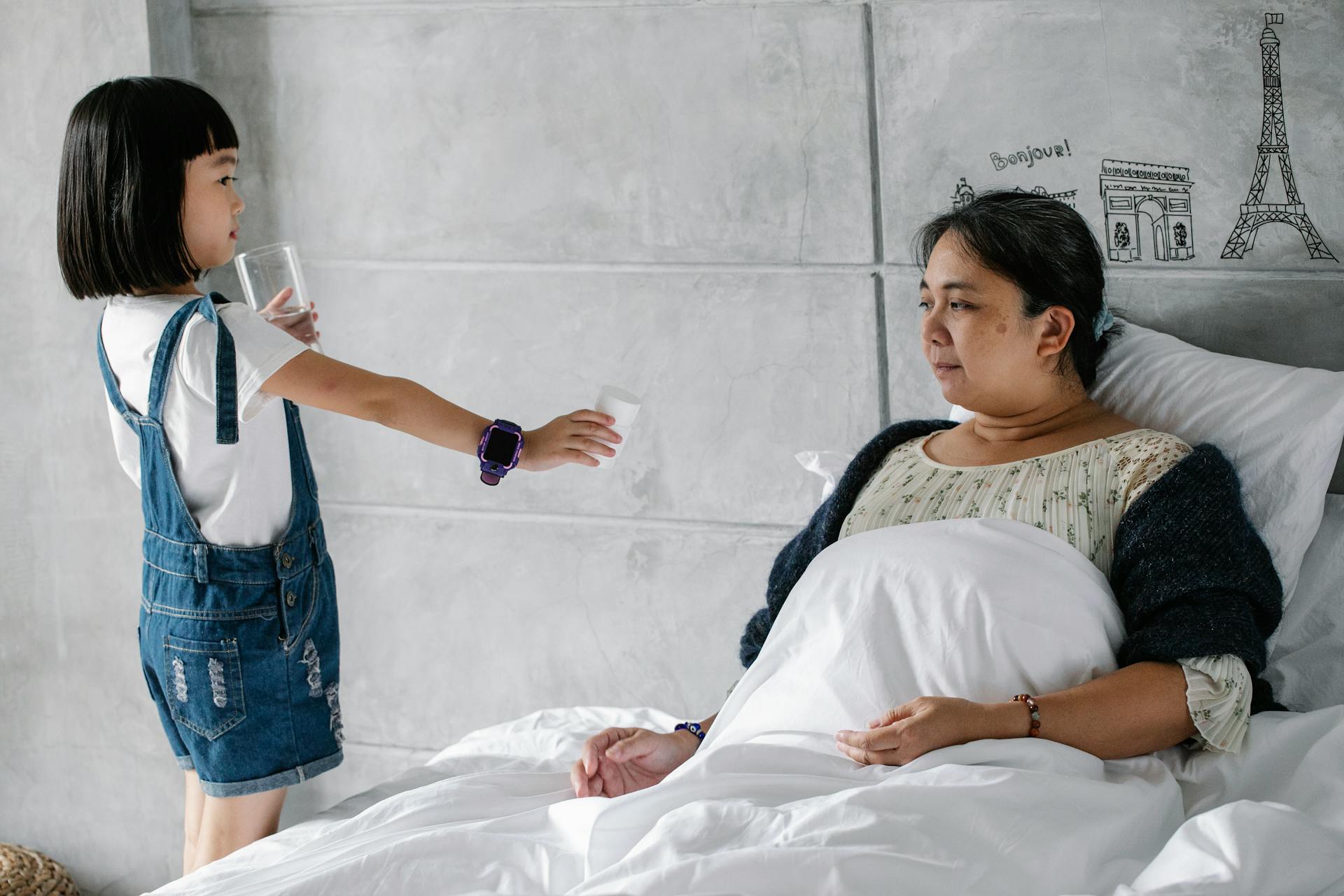 Side view of adorable little Asian girl giving medicine and water to sick grandmother sitting in bed