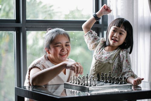 Веселая старшая этническая женщина с милой внучкой играет в шахматы дома