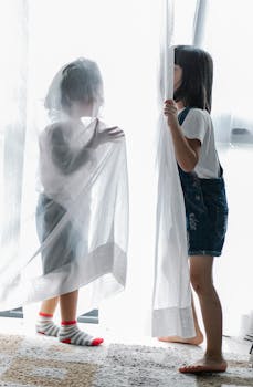 Side view of unrecognizable little siblings hiding behind curtains while playing together at home