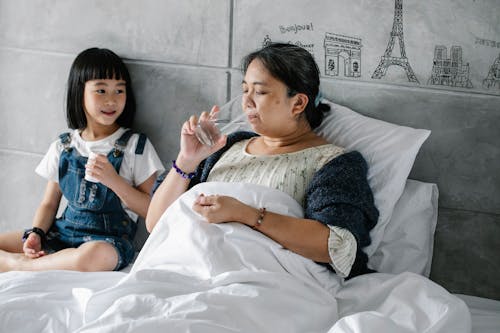 Adorable ethnic child giving pills to unhealthy grandmother drinking water in bed