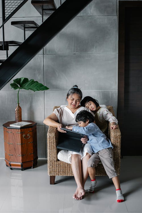Free Full body of happy Asian senior woman in casual clothes sitting in armchair with adorable little grandchildren and watching interesting video on tablet Stock Photo