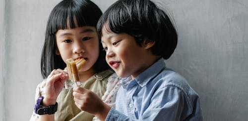 Adorable little Asian siblings with dark hair eating sweet yummy ice pop while standing near gray wall